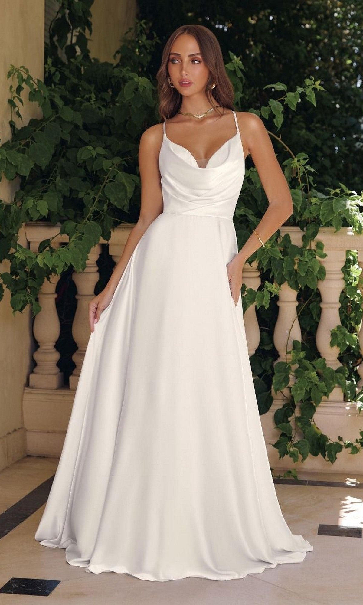 A woman in the Strappy-Back Long White Wedding Gown: Amoris T714W with a cowl neckline stands on a terrace, surrounded by lush greenery.